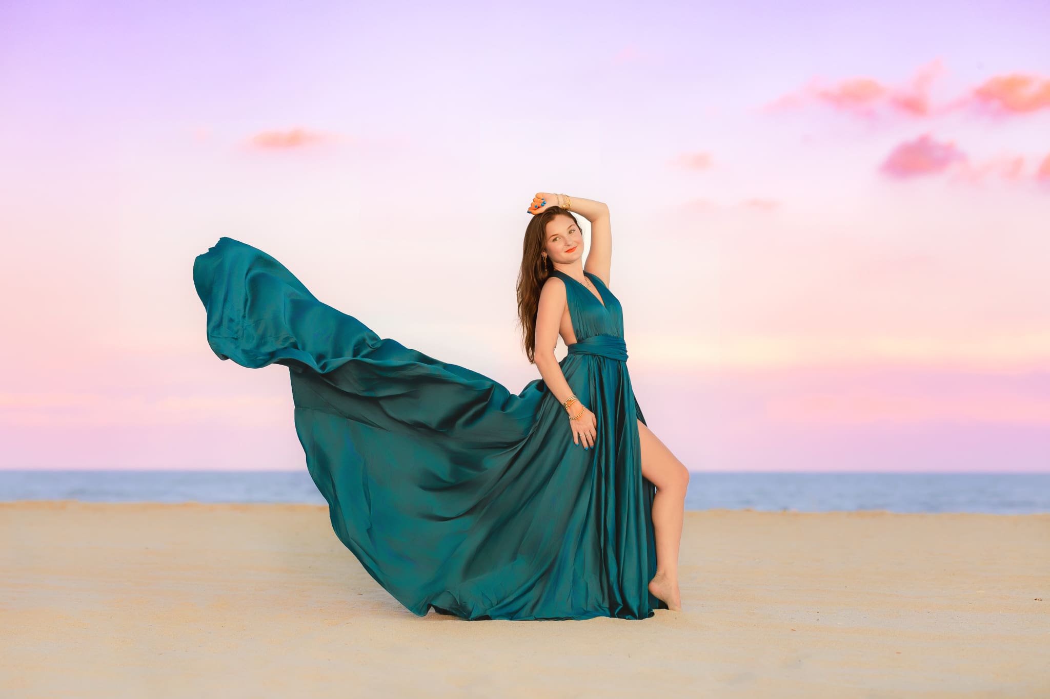 High School Senior posing on a beach for her senior photos in a gorgeous teal dress that is flowing backward away from her.