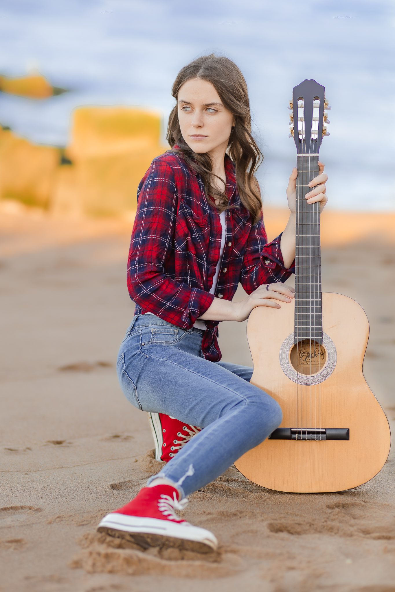 High school senior girl on a beach in a flannel shirt, jeans and red converse posing with her guitar for her senior photos.