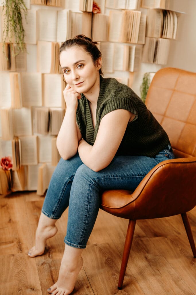 Female sitting on a chair in front of a stack of open books on the wall behind her for her senior photos.